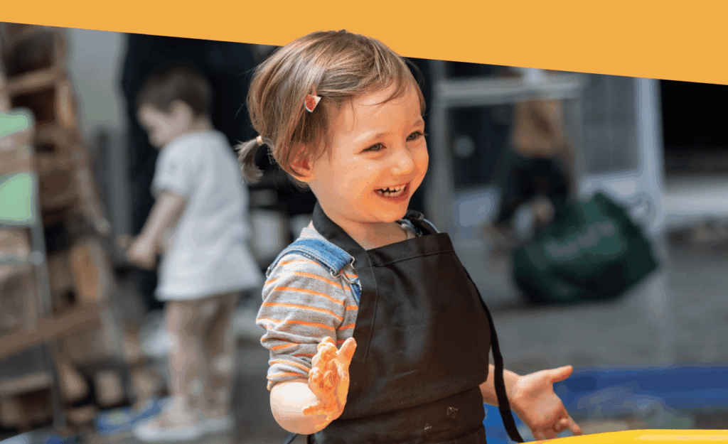 A joyful toddler in an apron, hands covered in paint, laughs while participating in a hands-on messy play art session.
